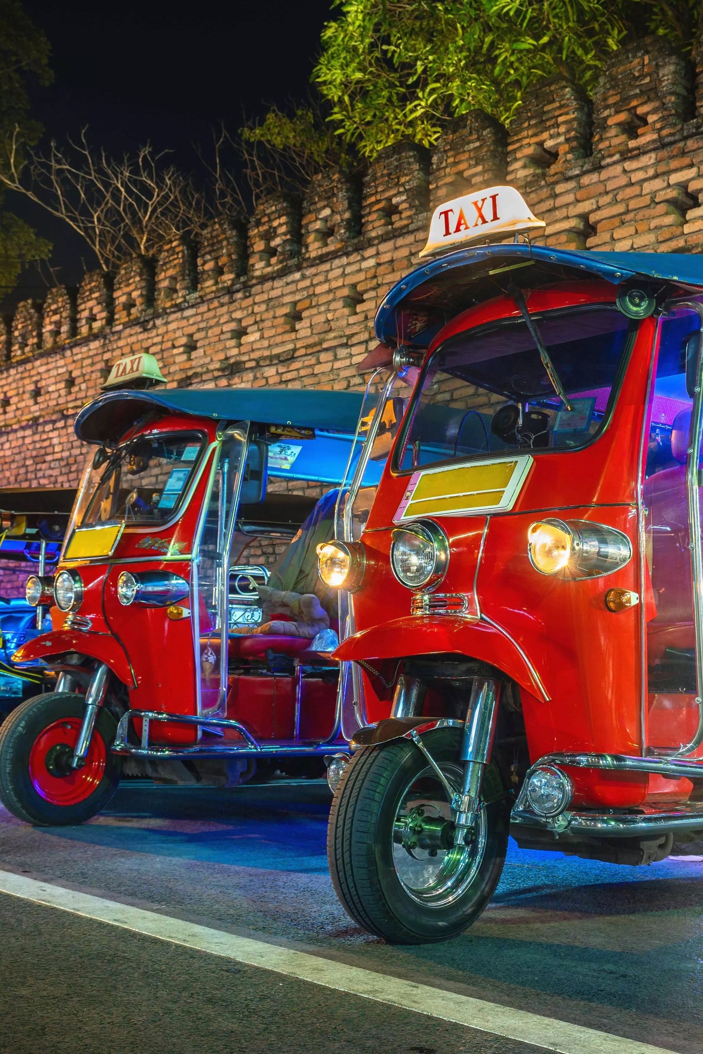Colorful tuk-tuks lined up against a brick wall at night.