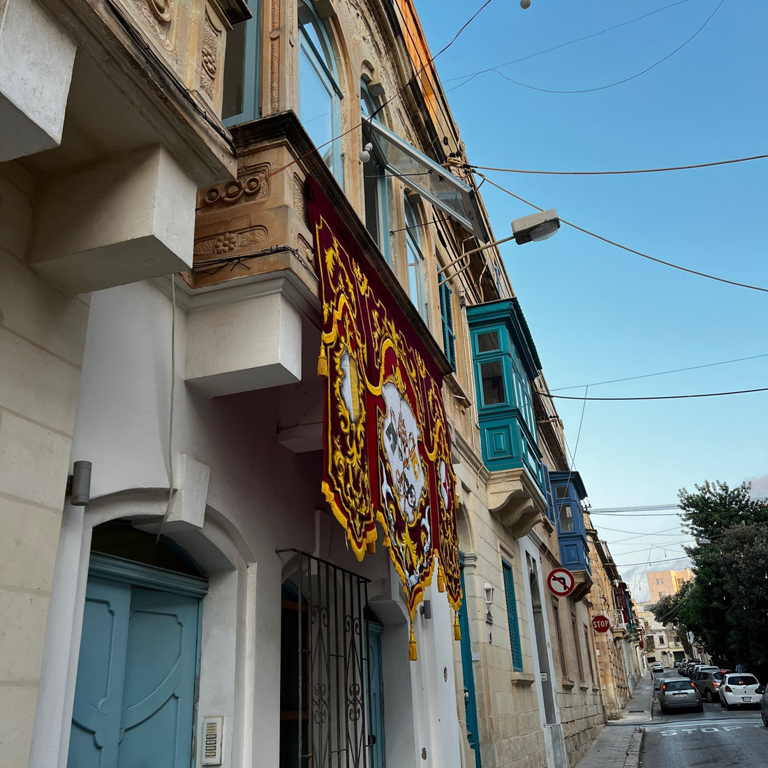 Ornate red and gold festa banner hanging over a narrow Maltese street lined with wooden balconies