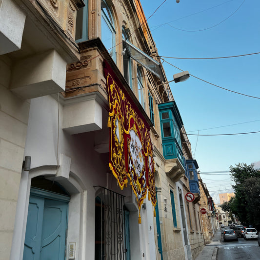 Ornate red and gold festa banner hanging over a narrow Maltese street lined with wooden balconies