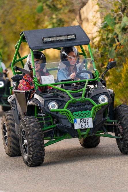 Green off-road vehicle with two people inside, driving on a road with trees in the background.