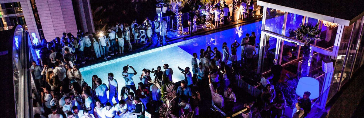 La azotea de Infinity by Hugo's de noche, en plena fiesta: la piscina infinita iluminada al borde del H Hotel, los jacuzzis, las palmeras y los sofás VIP bajo el techo abierto, con las luces de Paceville abajo