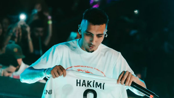 Person holding a sports jersey with 'Hakimi' printed on it, surrounded by people in a dark setting.
